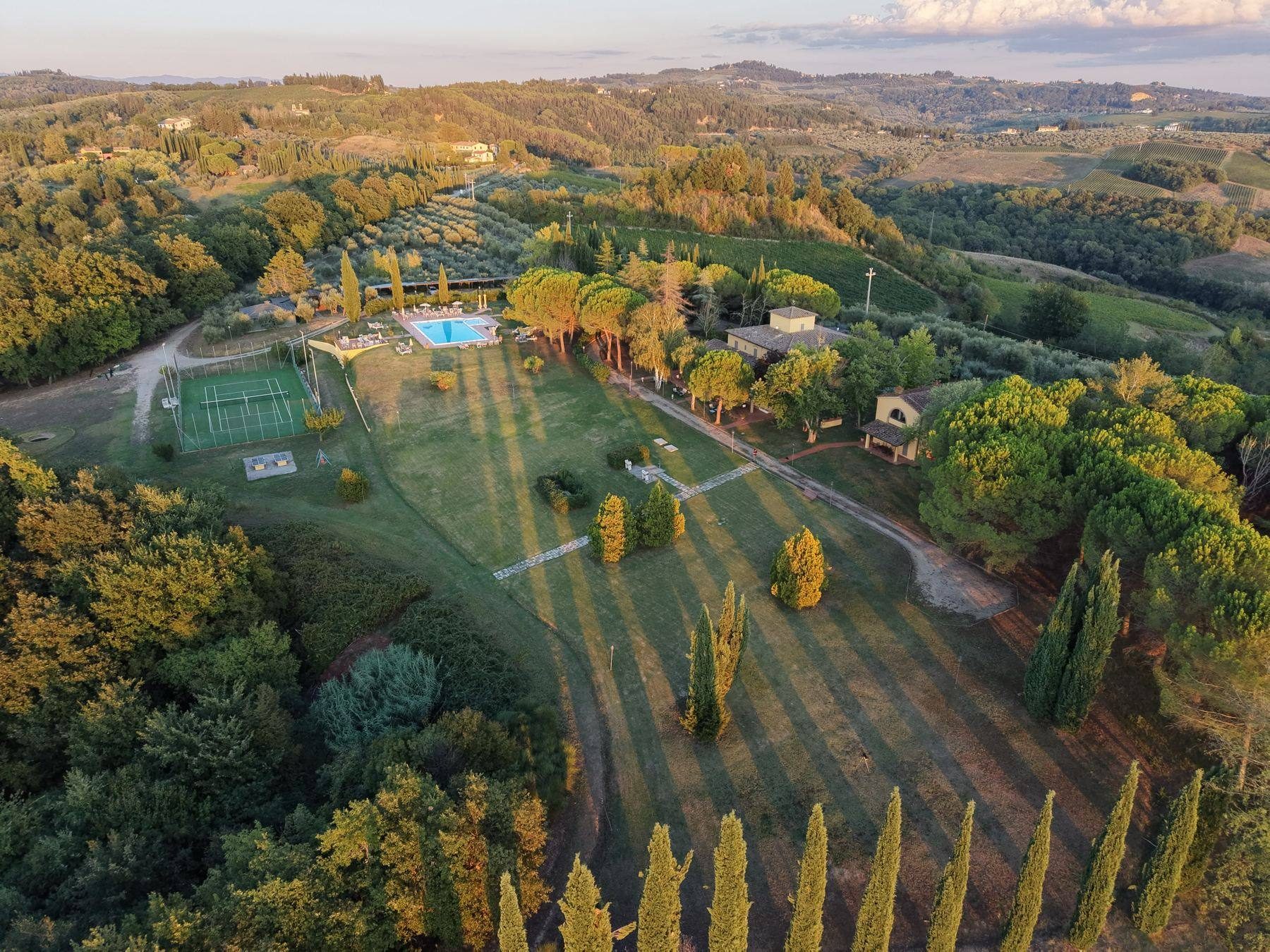 A private estate in the Tuscan hills from above, in golden evening light, with a pool, cypress trees and rolling hills
